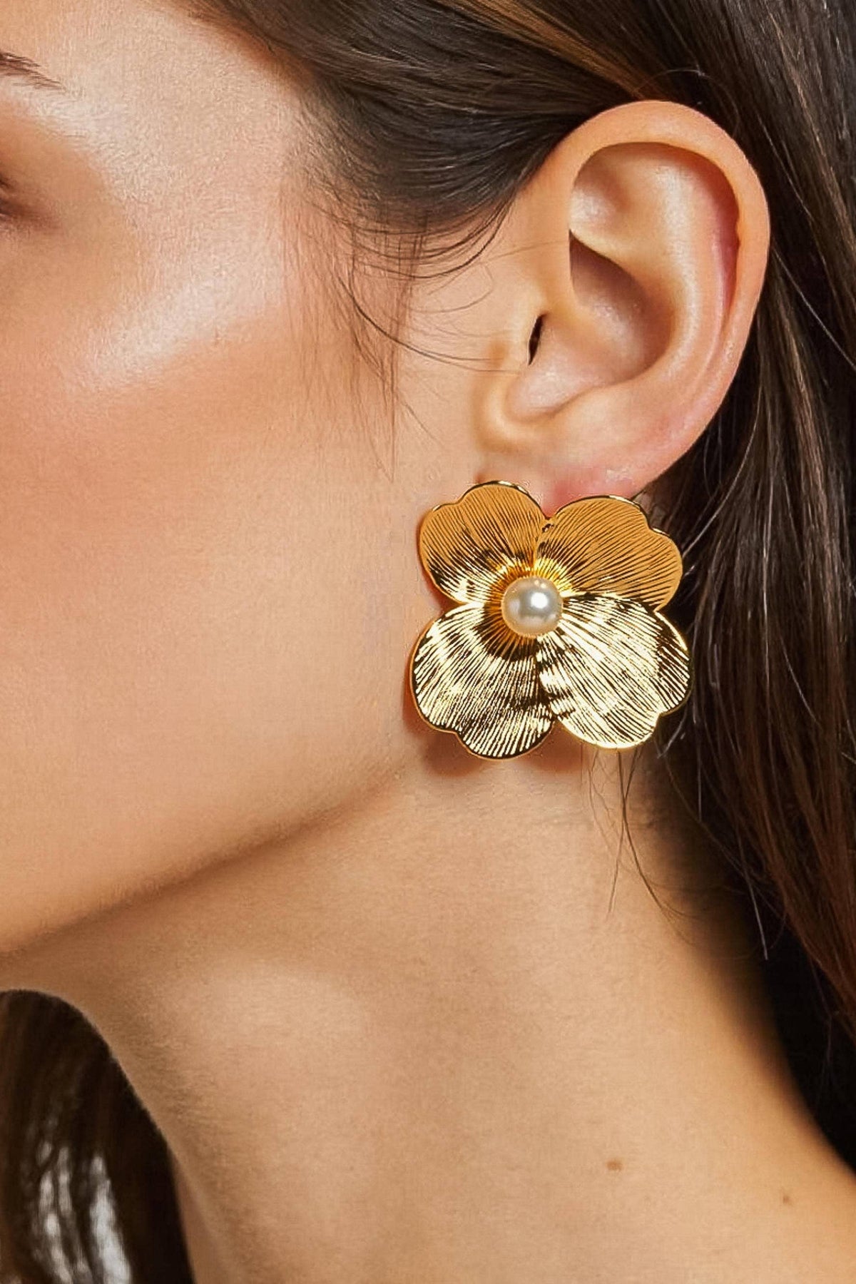 A close-up of a woman's ear wearing a textured gold flower-shaped earring with a small white pearl at the center.