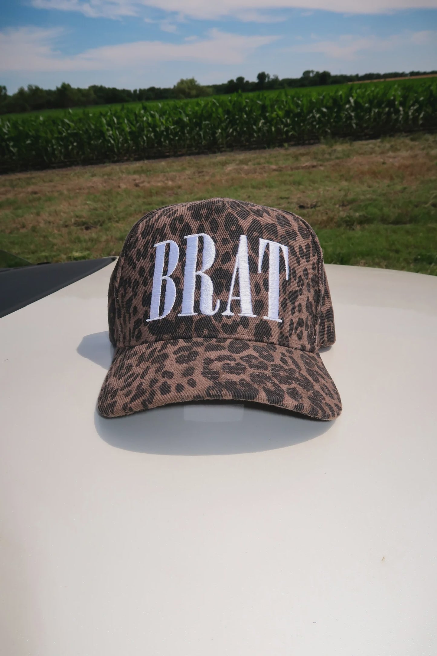 Leopard print cap with 'BRAT' text on a vehicle roof with a cornfield background