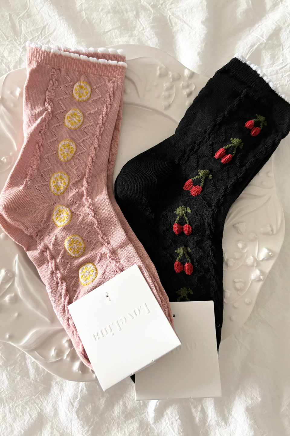 Two decorative crew socks laid flat on a white textured surface: one dusty pink sock with yellow lemon slice embroidery and one black sock with red strawberry embroidery, each with a white paper tag attached.