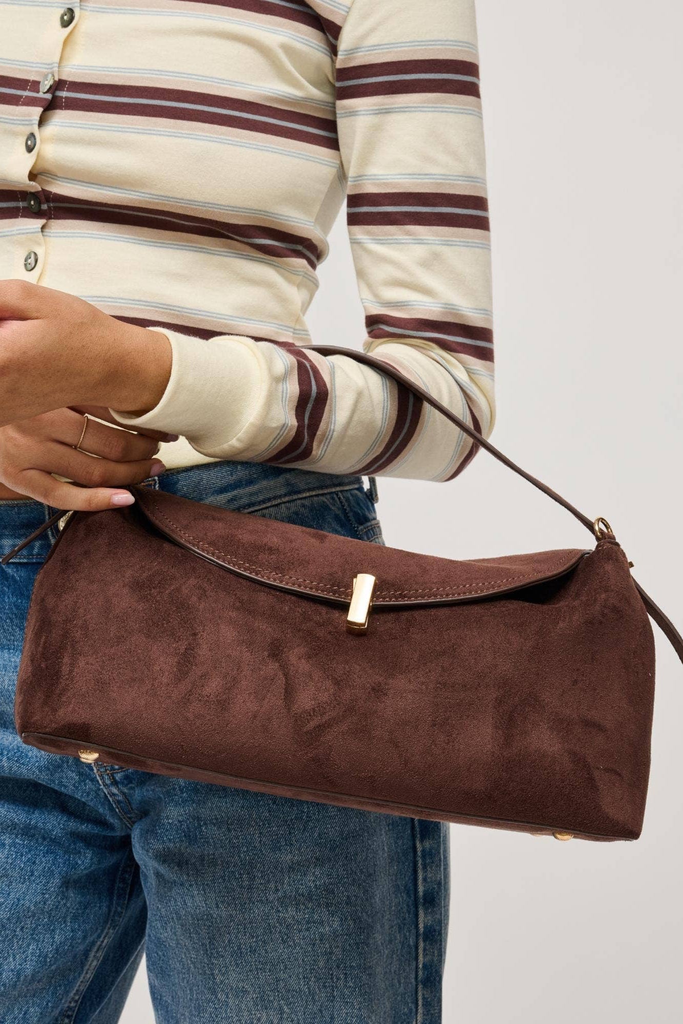 A person wearing a cream and brown striped long-sleeve top and blue jeans holds a brown suede shoulder bag with a gold-tone clasp against a light gray studio background.