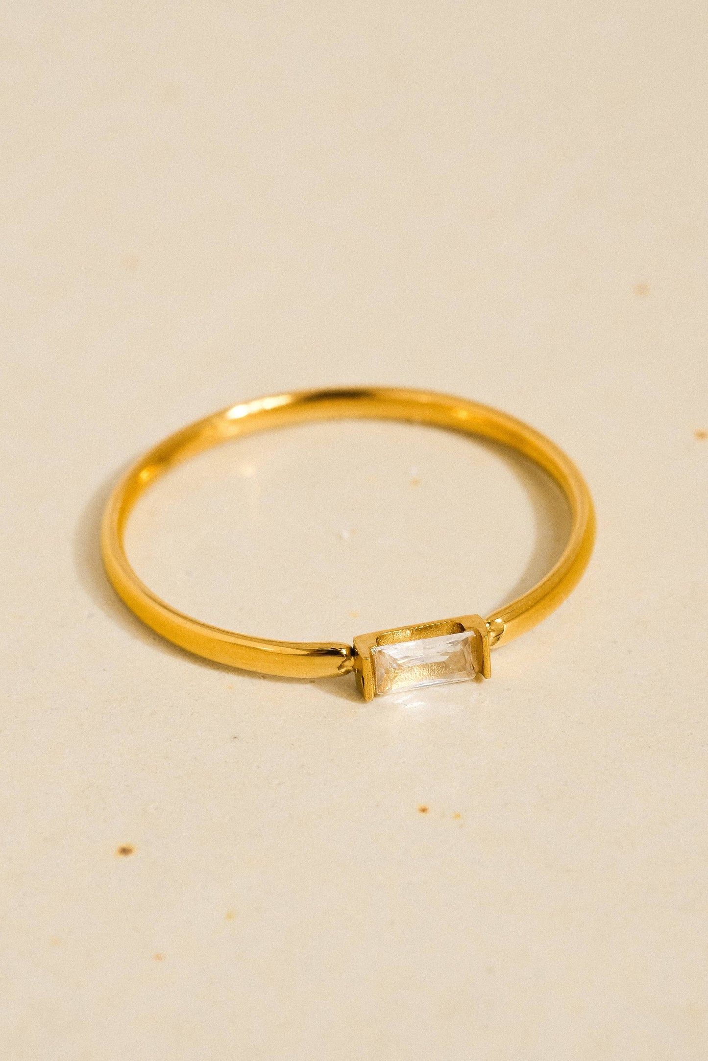 A gold ring with a rectangular clear gemstone, photographed on a light beige textured surface.