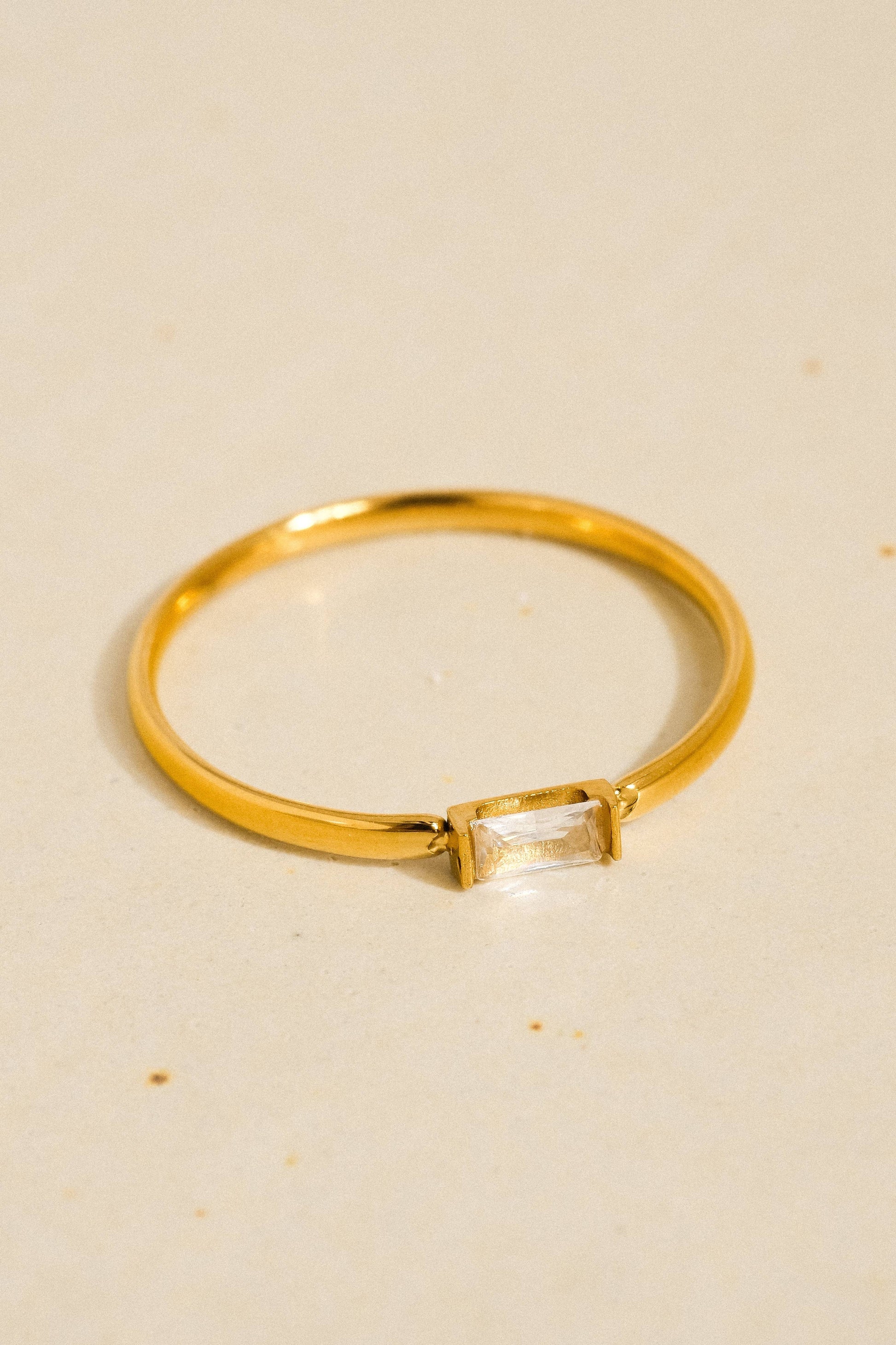 A gold ring with a rectangular clear gemstone, photographed on a light beige textured surface.