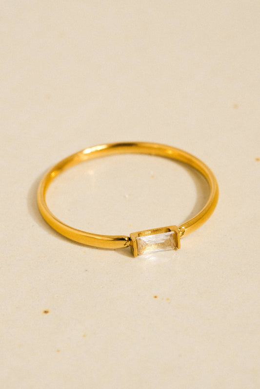 A gold ring with a rectangular clear gemstone, photographed on a light beige textured surface.