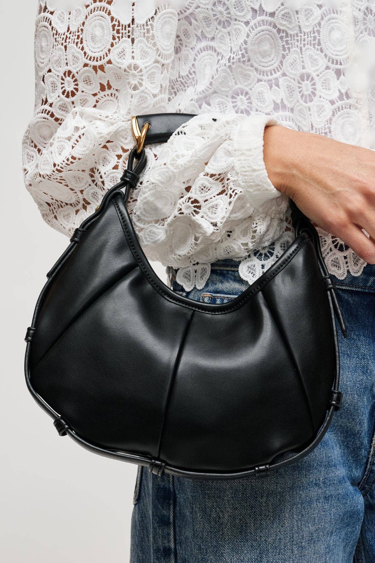 A person wearing a white lace blouse and blue jeans carries a black crescent-shaped shoulder bag with gold-tone hardware against a light gray background.