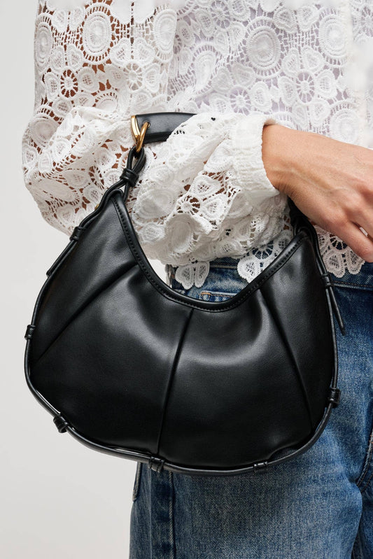 A person wearing a white lace blouse and blue jeans carries a black crescent-shaped shoulder bag with gold-tone hardware against a light gray background.