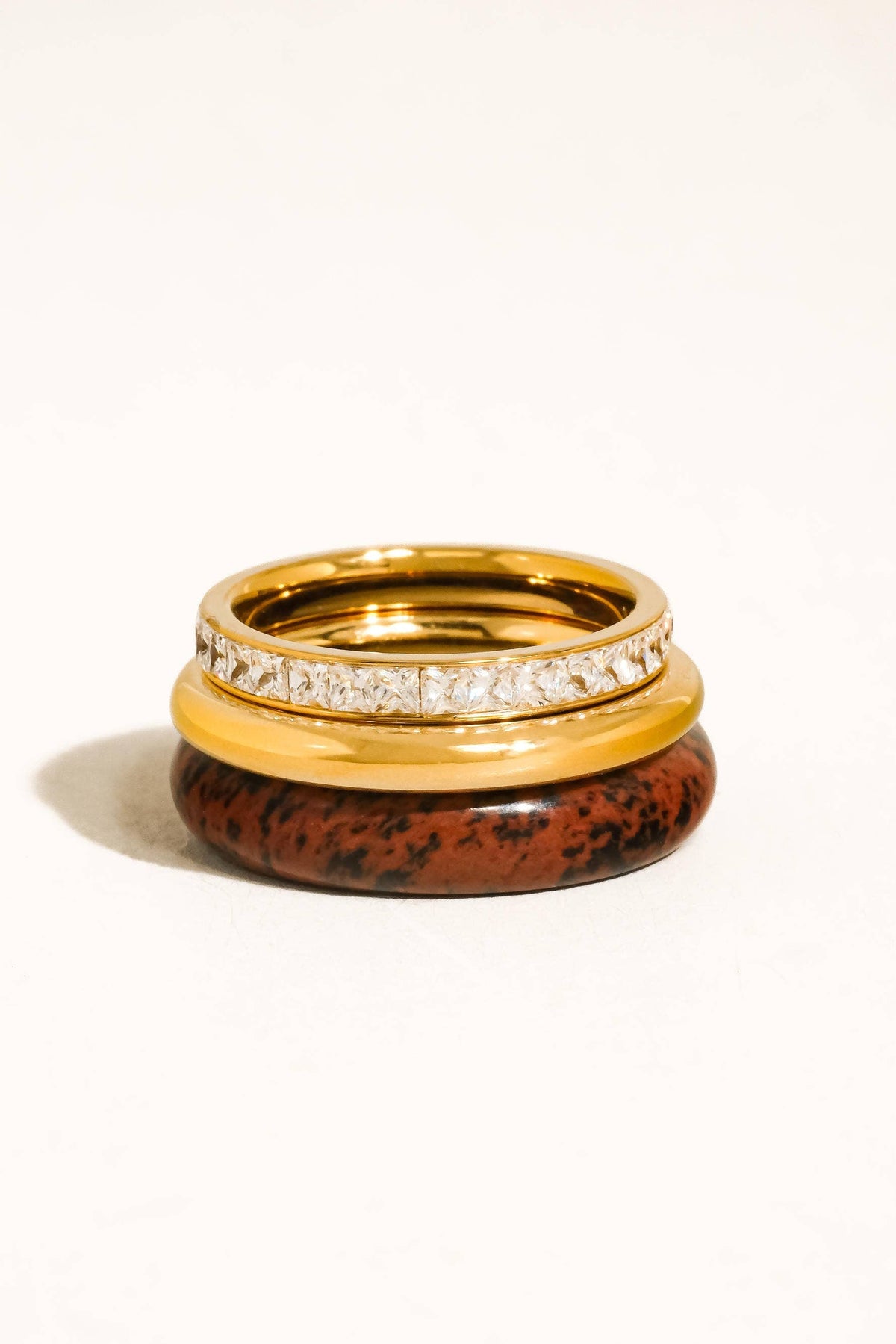 A stack of three rings on a plain white background. The top ring is a polished yellow gold band with a row of clear stones.