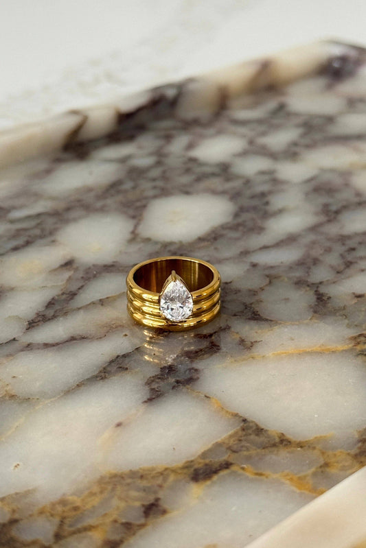A gold ring with a pear-cut clear gemstone, displayed on a polished white and brown marble tray.
