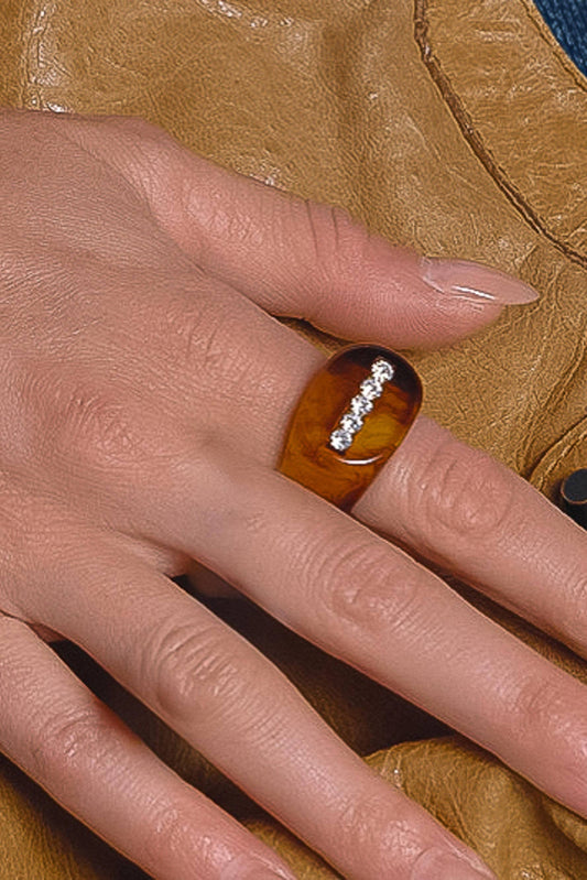 A close-up of a hand wearing a chunky amber-brown ring with a row of small clear stones, resting against a tan leather surface.