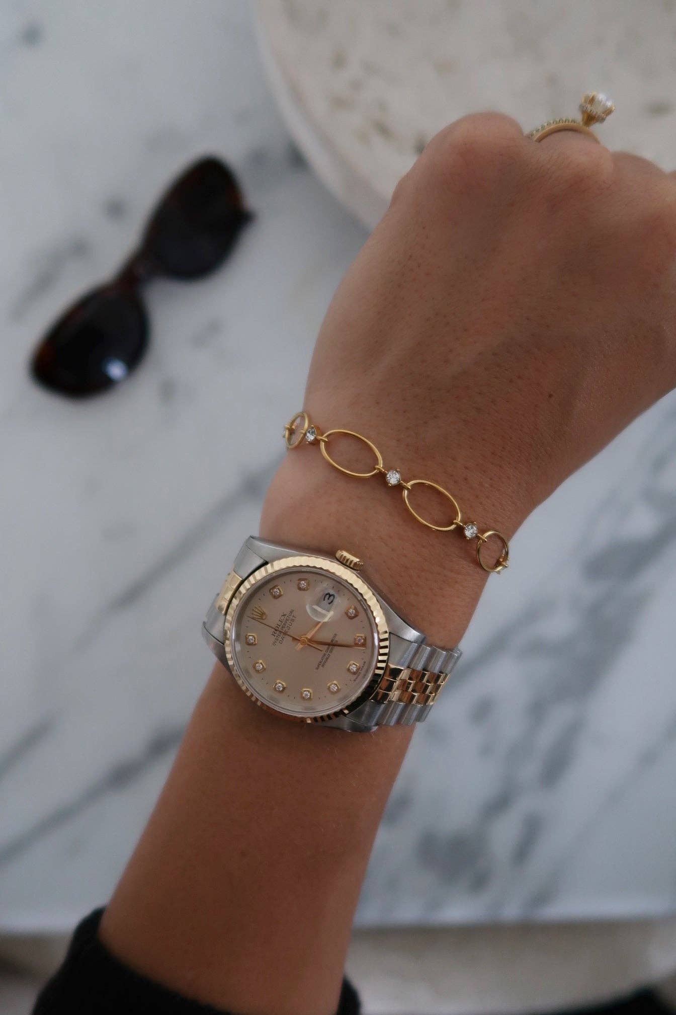 Hand wearing a gold watch and bracelet on a marble surface with sunglasses.