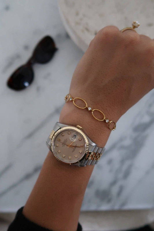Hand wearing a gold watch and bracelet on a marble surface with sunglasses.