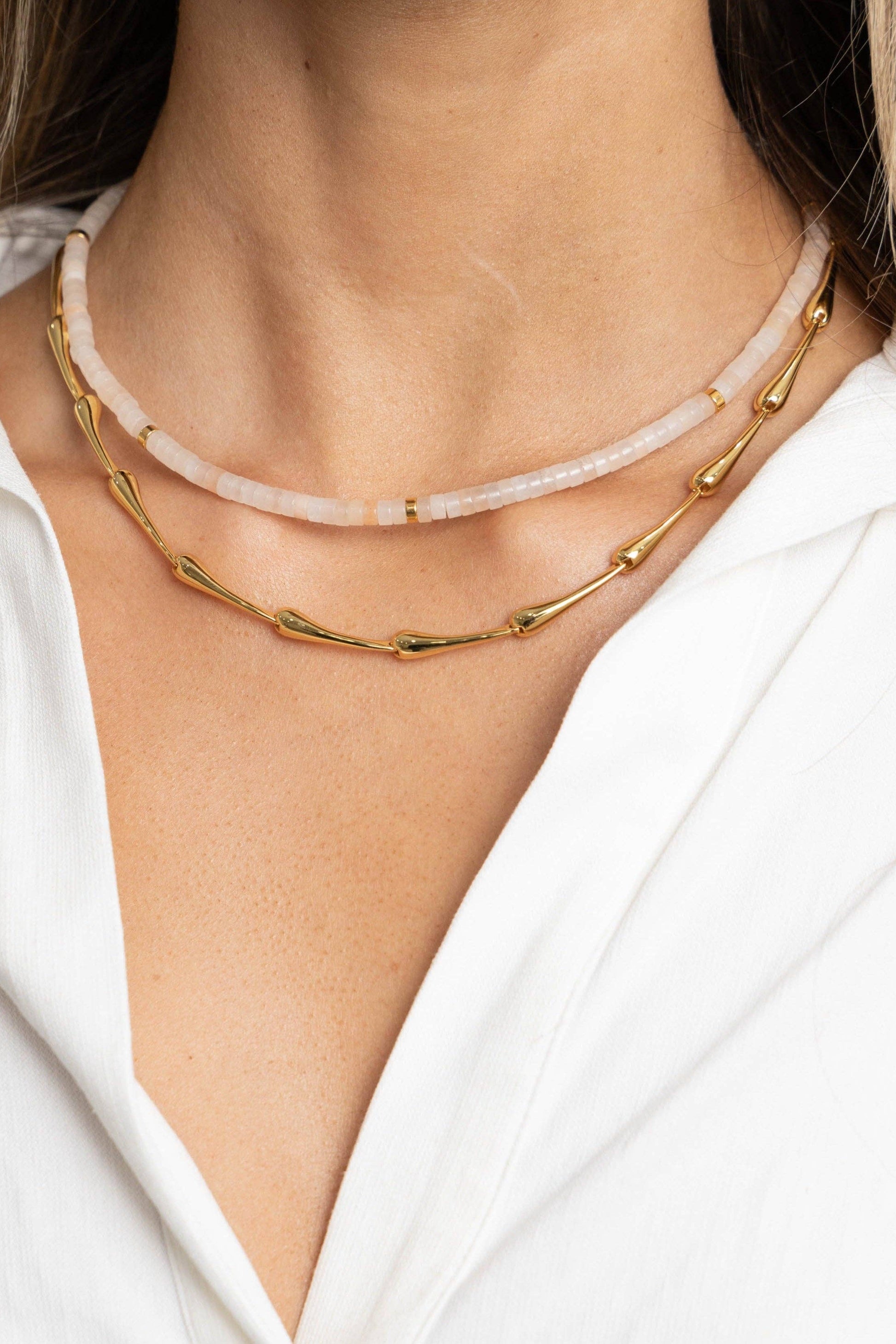 Two necklaces, one gold and one pink, on a person wearing a white shirt.