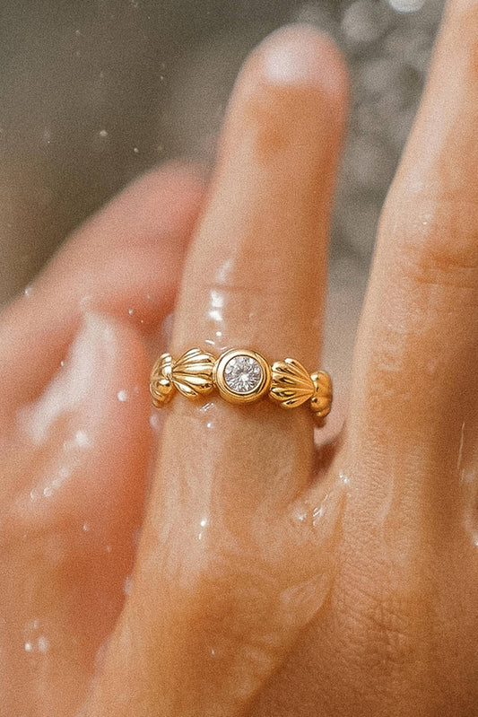 A gold ring with a central round clear gemstone and decorative shell-like side details, shown worn on a finger with water droplets in the foreground.
