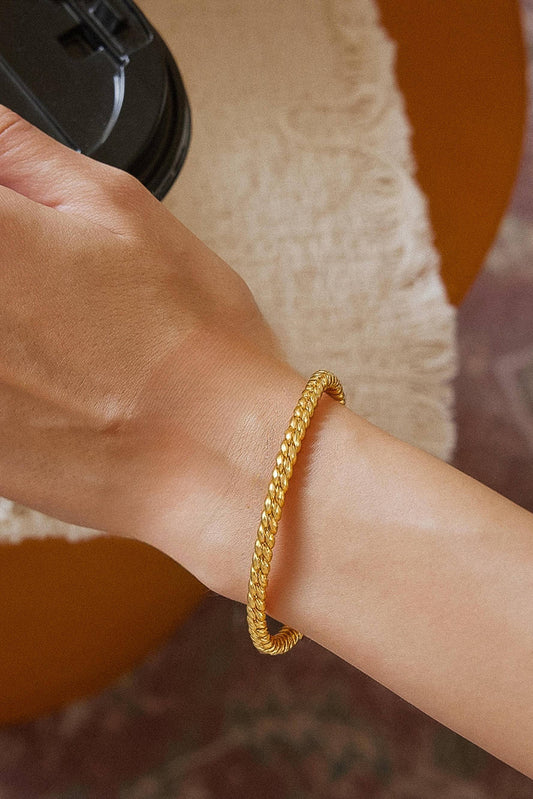 A close-up of a person's wrist wearing a textured gold bangle bracelet, with a beige fringed fabric and an orange-brown circular background visible.