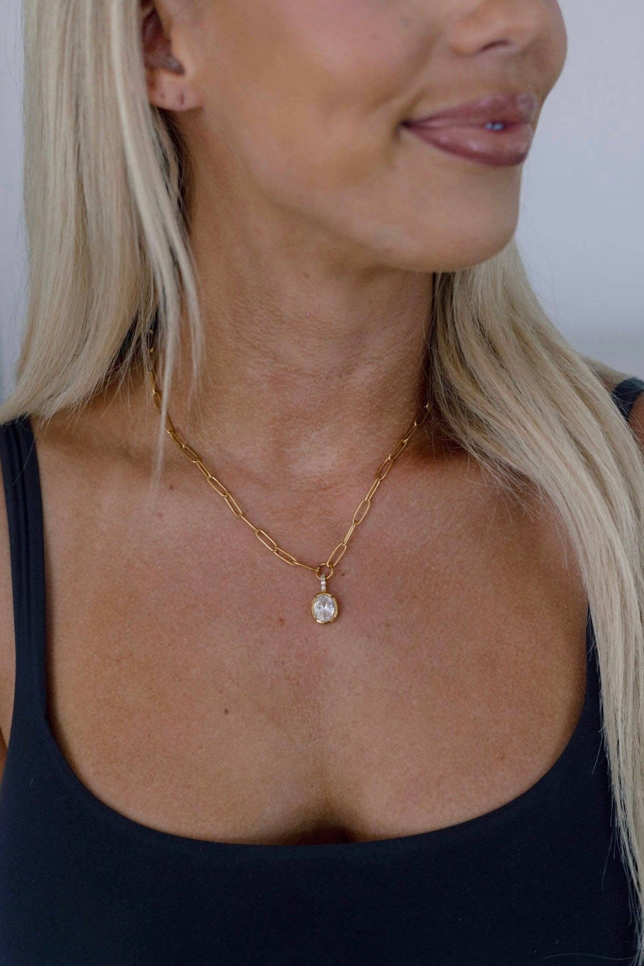 A close-up of a blonde woman wearing a delicate gold chain necklace with a small, clear oval pendant.