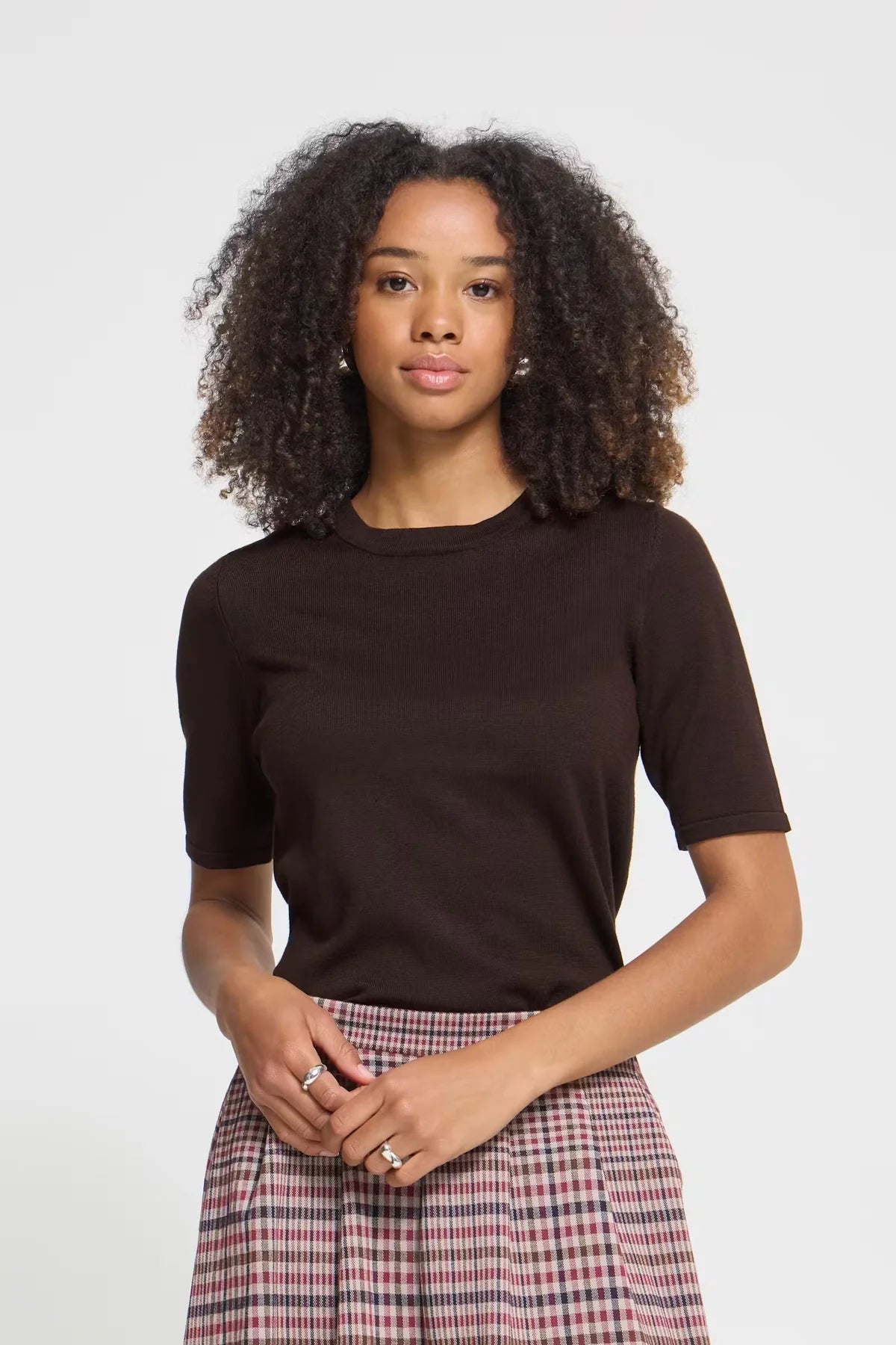 A woman with curly dark hair wearing a dark brown short-sleeve crewneck top and a pink, red, and white plaid skirt, standing against a light gray studio background.