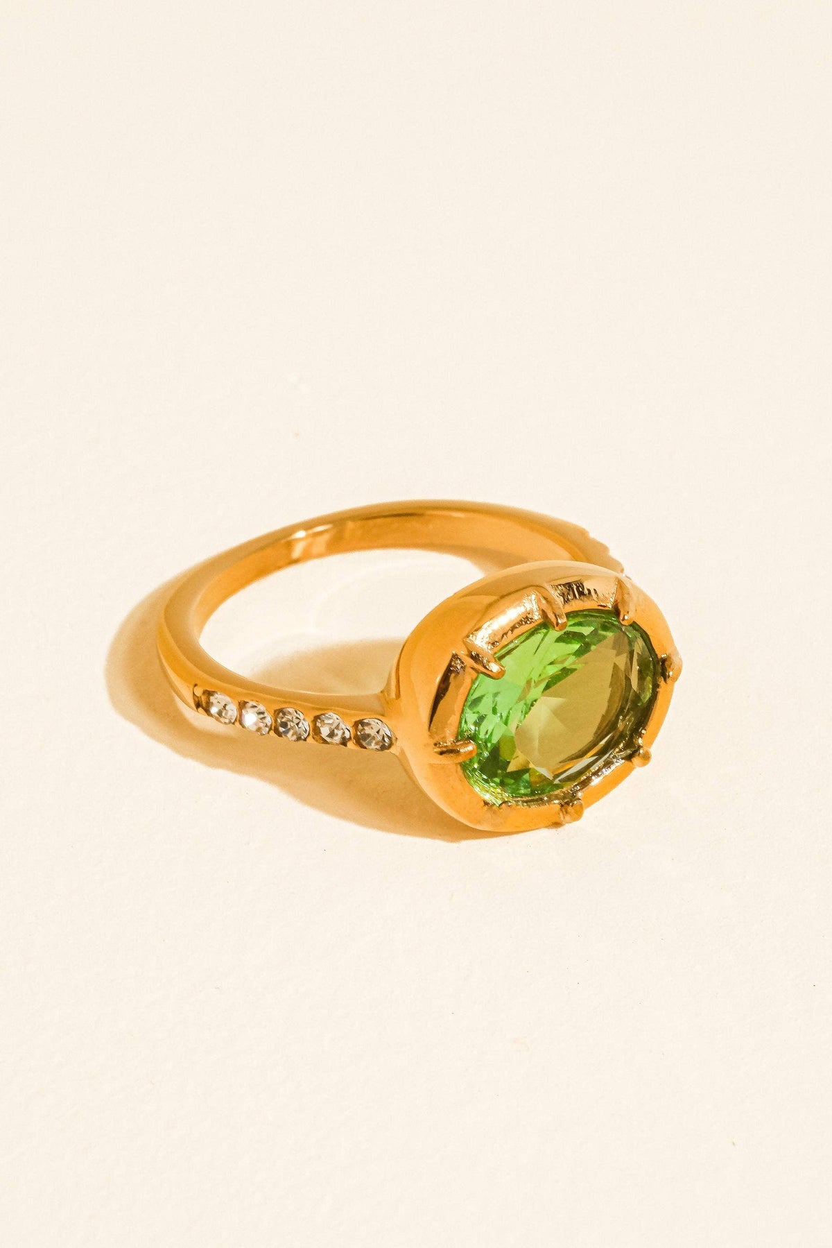 A gold ring featuring a large oval green gemstone and a row of small clear stones on the band, set against a plain off-white background.