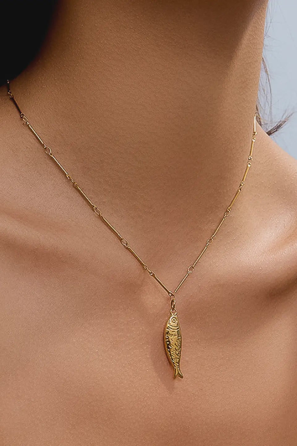 Gold necklace with a fish pendant worn by a person against a light blue background