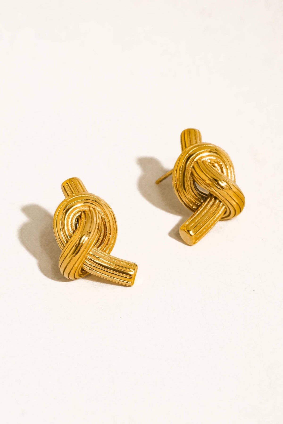 A pair of gold knotted post earrings with a textured, ribbed finish, displayed on a clean off-white background with soft shadows.