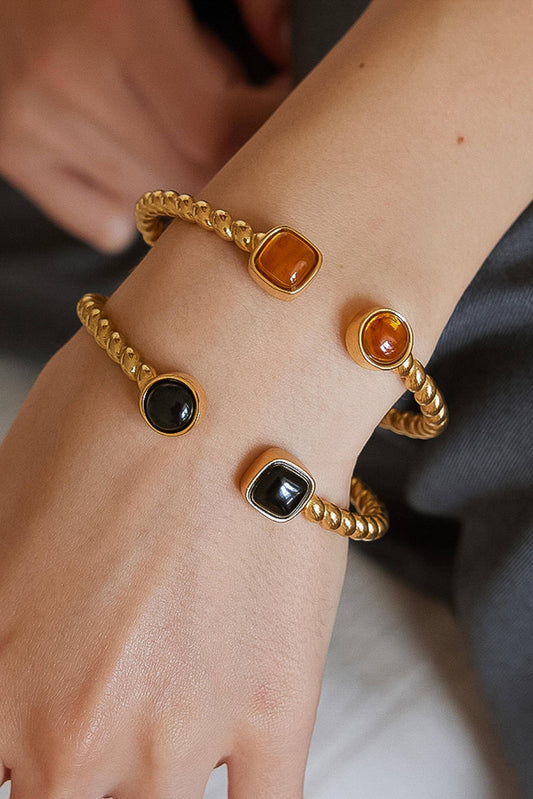 A close-up of a person's wrist wearing four gold-toned twisted bangle bracelets with square and round cabochon stones in black and amber-orange tones.