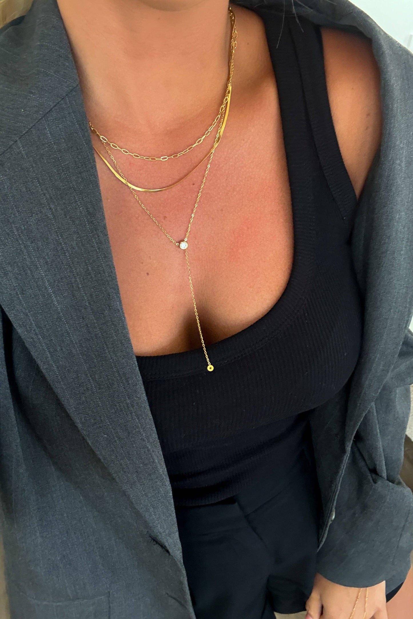 A close-up of a person wearing layered gold necklaces over a black ribbed tank top and a gray blazer.