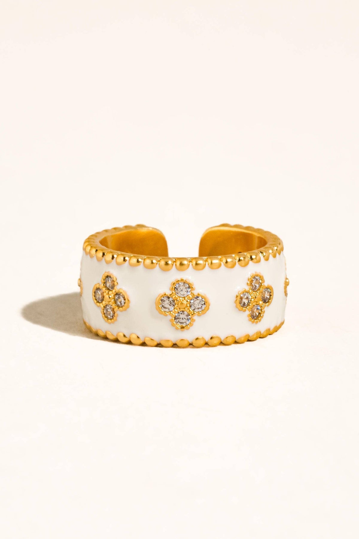 A wide white cuff bracelet with gold-tone beaded edges and three clover motifs paved with small clear stones, shown on a warm off-white background.