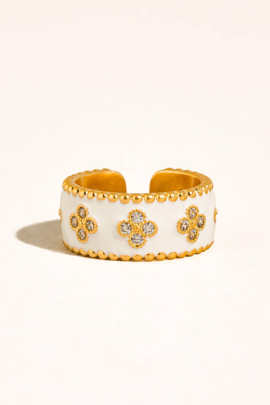 A wide white cuff bracelet with gold-tone beaded edges and three clover motifs paved with small clear stones, shown on a warm off-white background.