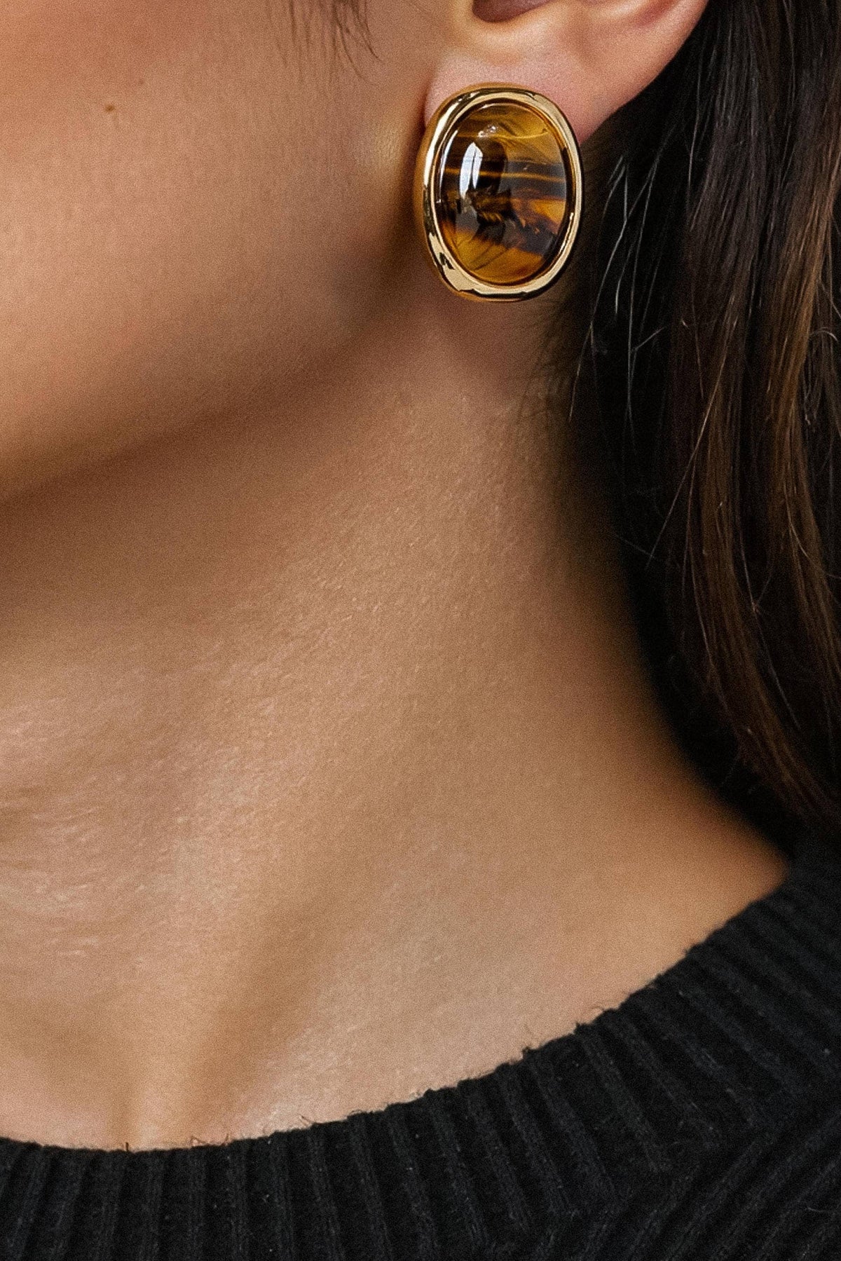 A close-up of a model's ear wearing a gold oval stud earring with a brown tiger's eye stone.