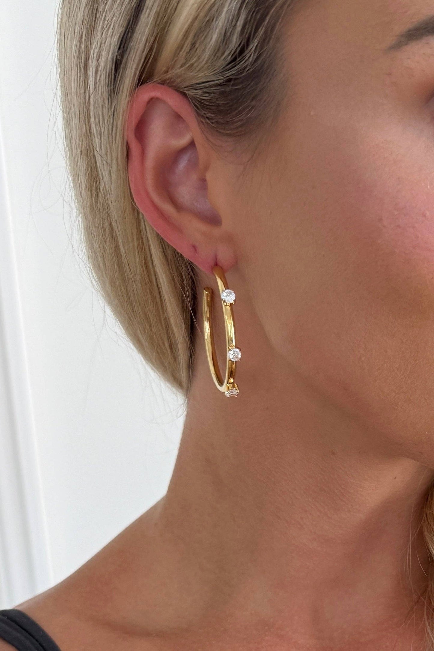 A close-up of a woman's ear wearing a pair of gold hoop earrings with small clear gemstone accents, against a light neutral background.