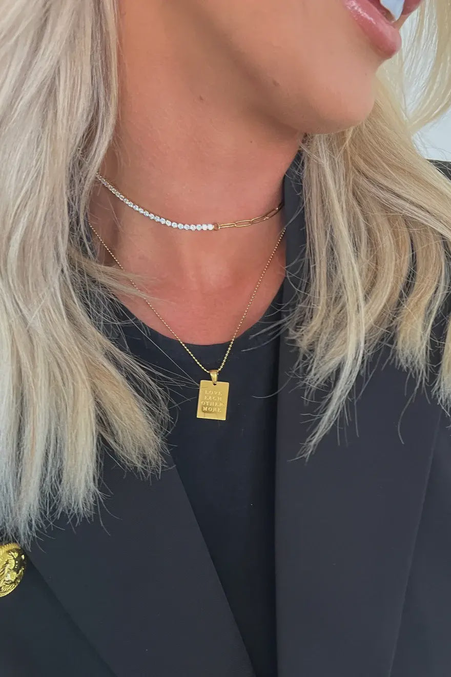Close-up of a person wearing a gold necklace with a rectangular pendant, against a neutral background.