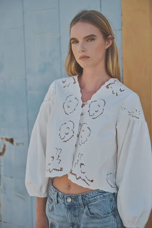 Woman wearing a white embroidered blouse and blue jeans against a rustic wall.