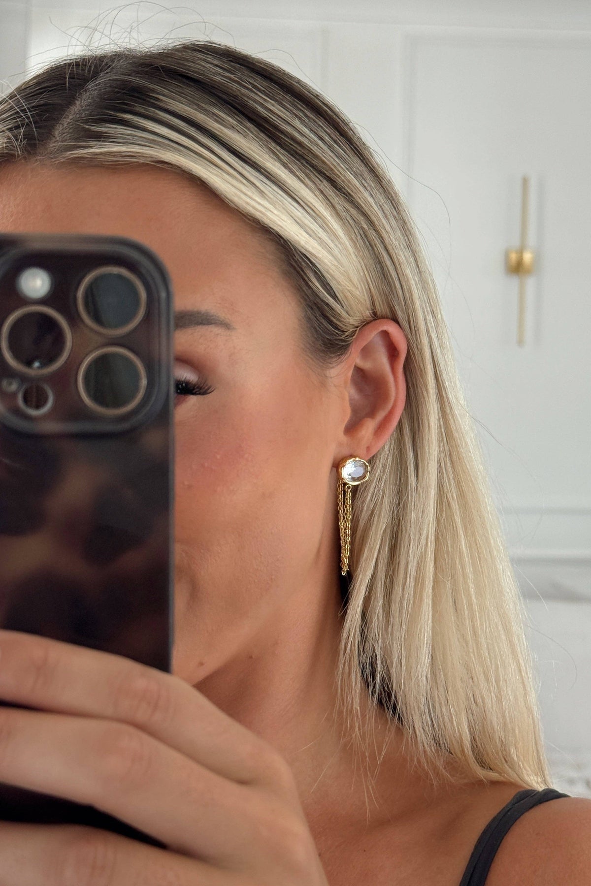 A blonde woman taking a mirror selfie to showcase a long, dangling gold earring with a clear round gemstone and small gold leaf accents.