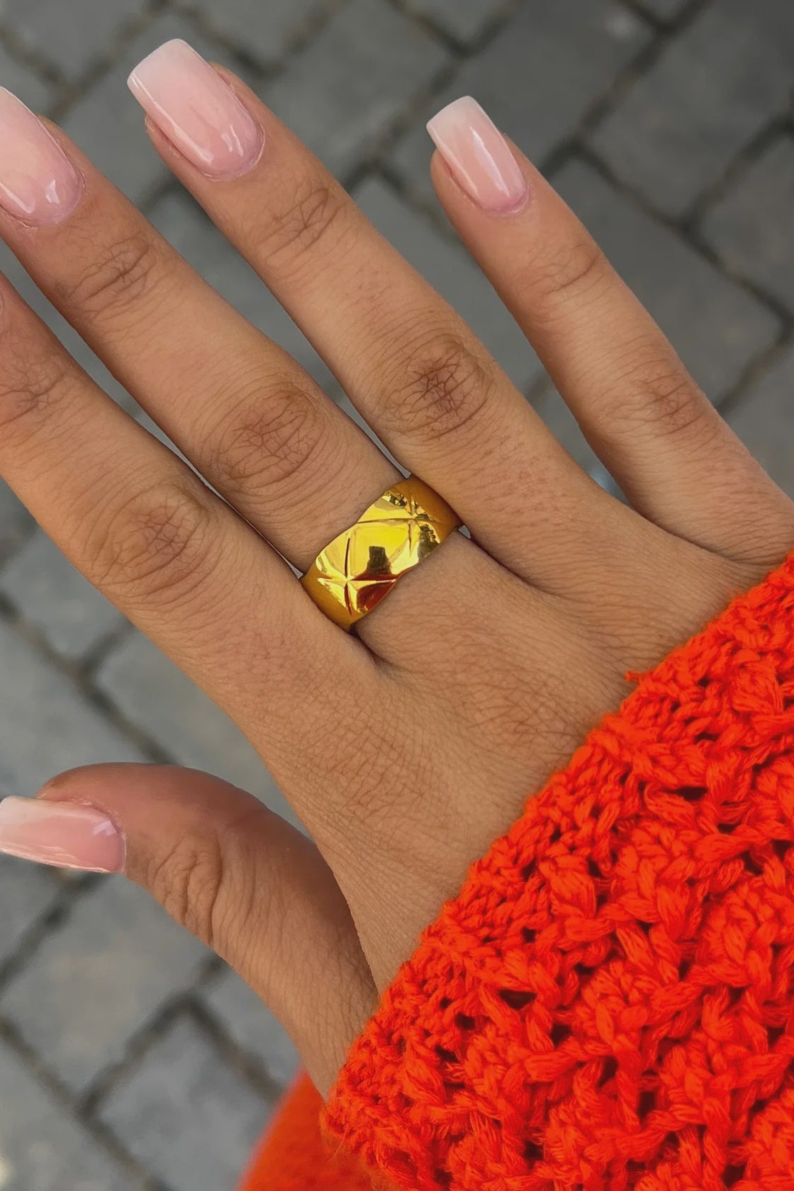 Hand wearing a gold ring with pink nail polish against a textured background