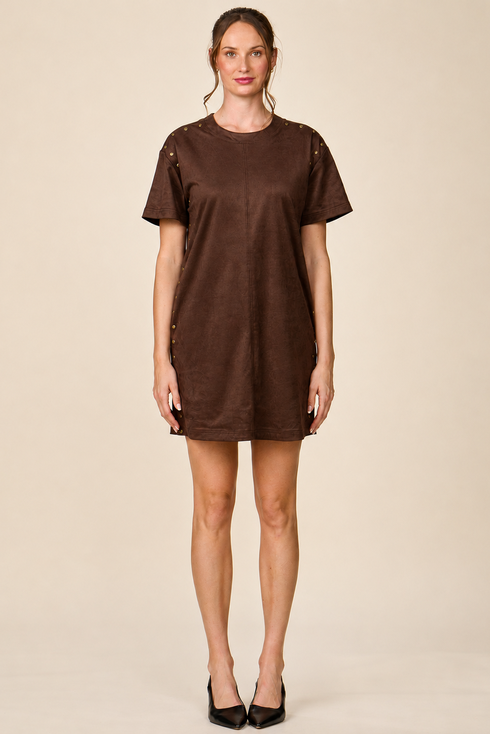 A woman modeling a brown short-sleeve t-shirt dress with decorative button details on the shoulders and sides, paired with black heels, against a solid beige background.