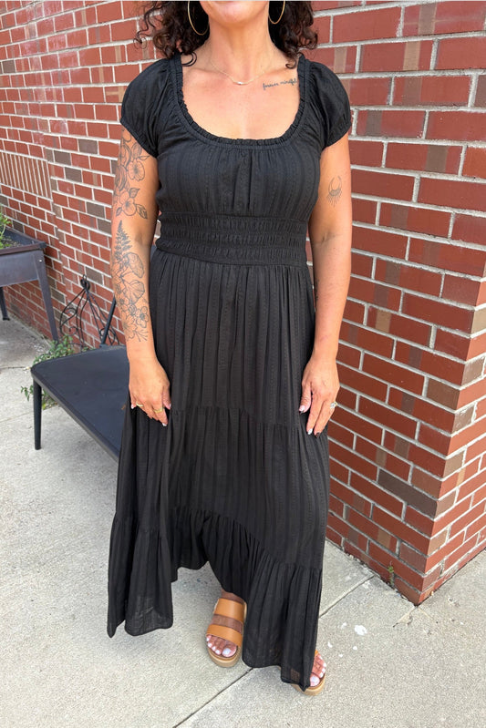 A woman wearing a black tiered maxi dress with short puff sleeves and a smocked bodice, standing outdoors against a red brick wall.