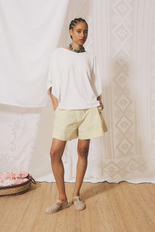 Woman wearing a white top and yellow shorts standing against a textured white curtain.