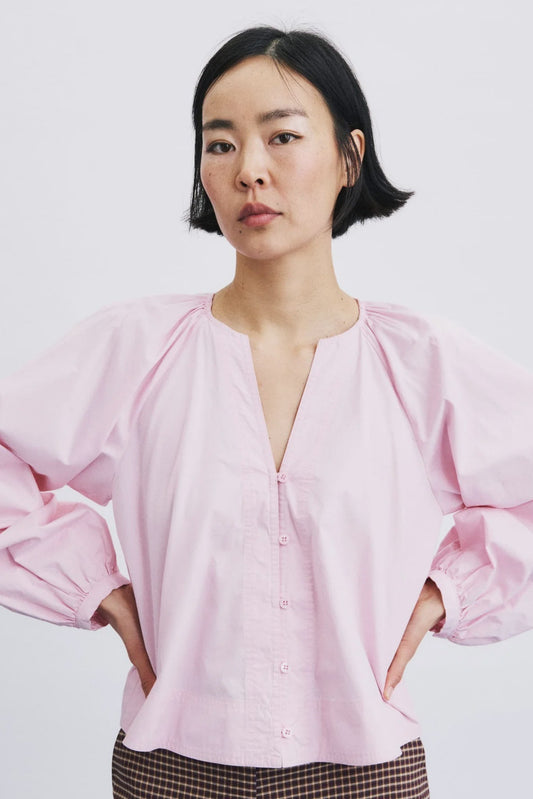 A woman wearing a light pink blouse with voluminous long sleeves, a split neckline, and front buttons, paired with brown and black checkered trousers, against a plain white background.