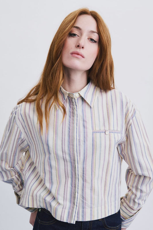 A woman with long red hair wearing a long-sleeve button-up shirt with vertical stripes in cream, blue, and purple, paired with dark jeans, against a plain white background.