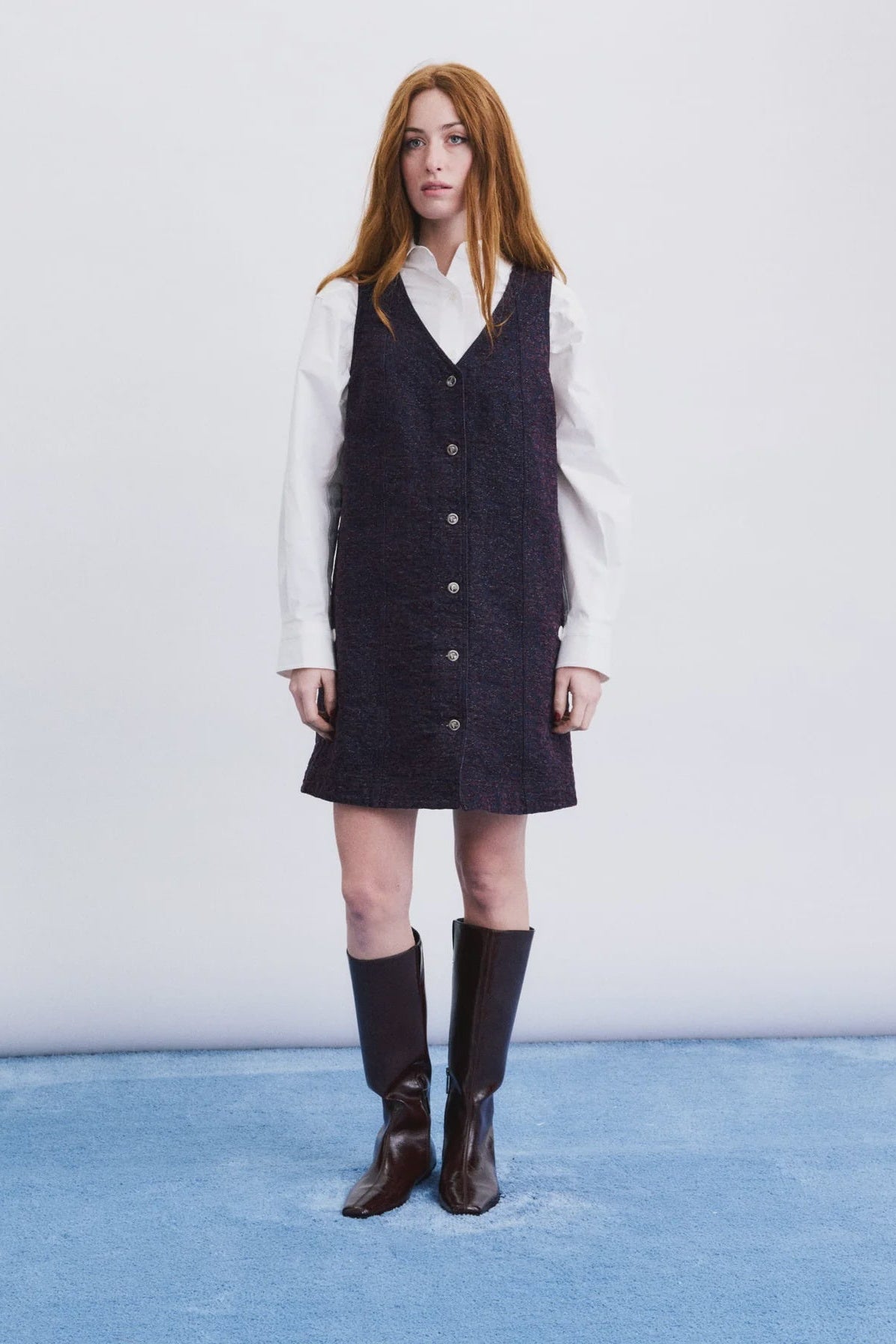 A model stands against a white studio wall wearing a dark navy textured button-front vest over a white collared shirt, paired with dark brown knee-high boots.
