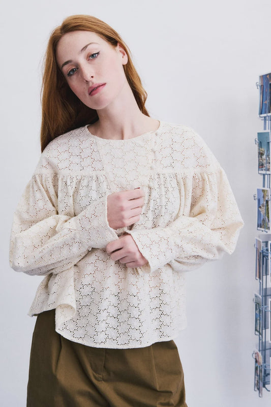 A woman wearing a cream-colored, long-sleeved lace blouse with a gathered ruffle at the neckline, paired with olive green pants, standing against a white wall next to a wall-mounted photo rack.
