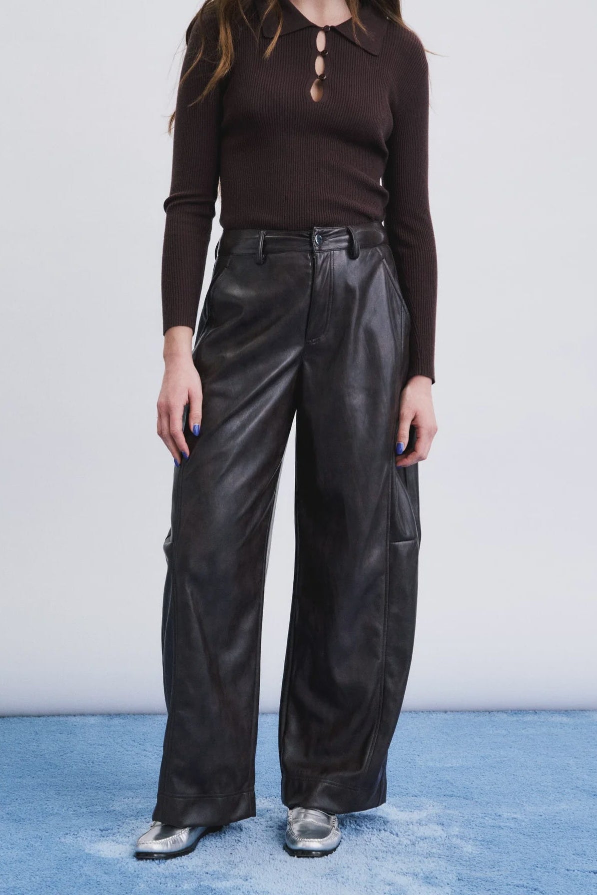 A model wearing a dark brown long-sleeve polo shirt and black wide-leg leather pants, standing against a white wall with a blue textured floor.