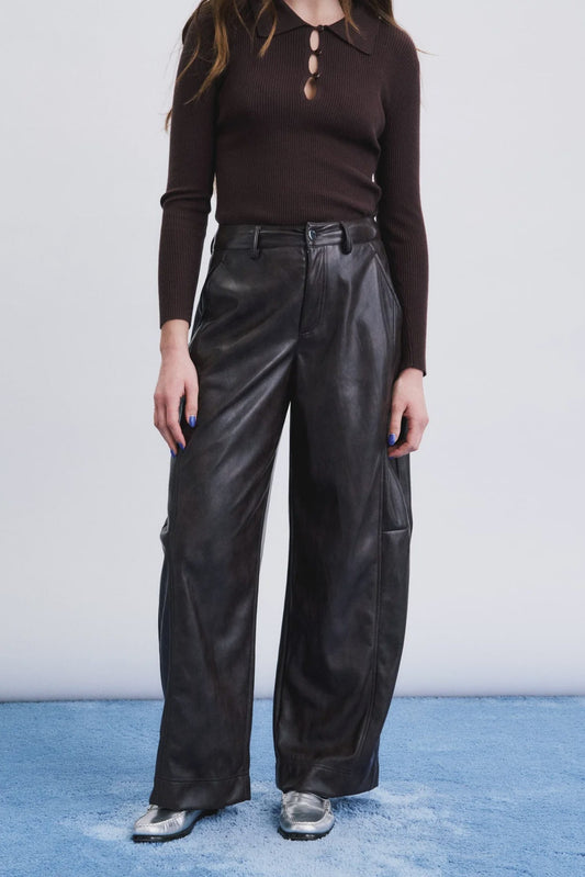 A model wearing a dark brown long-sleeve polo shirt and black wide-leg leather pants, standing against a white wall with a blue textured floor.
