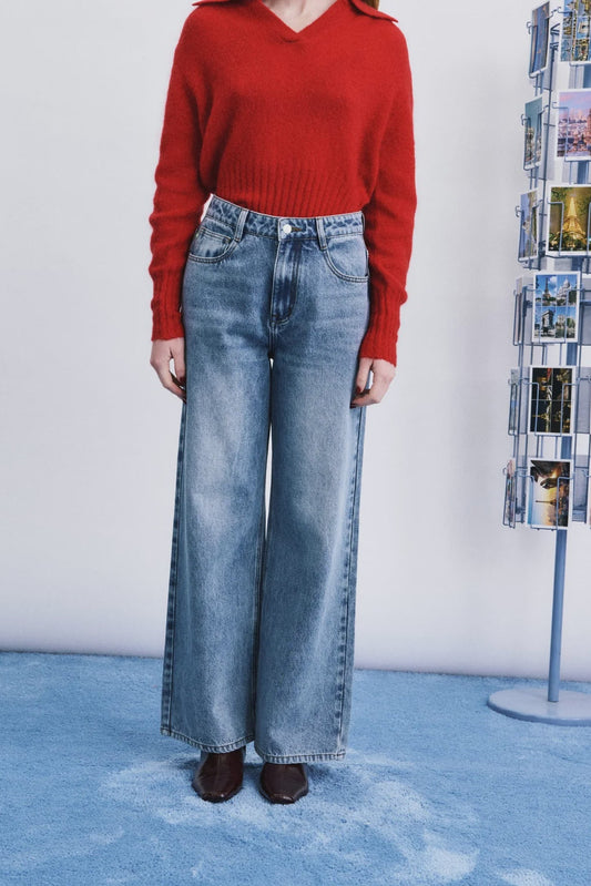 A model wearing a vibrant red knit sweater and high-waisted, wide-leg blue denim jeans, standing against a white wall with a blue textured rug and a magazine rack in the background.