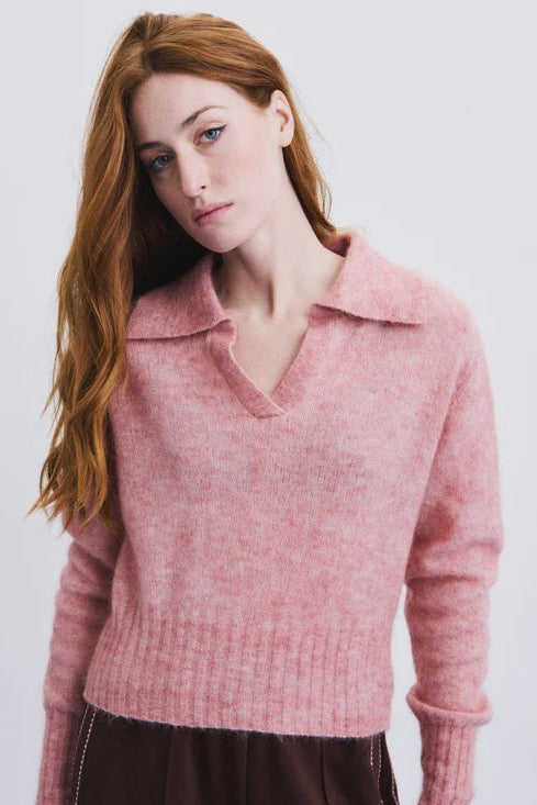 A woman with long red hair wearing a soft pink knit polo sweater and a dark brown skirt, photographed against a clean white studio background.