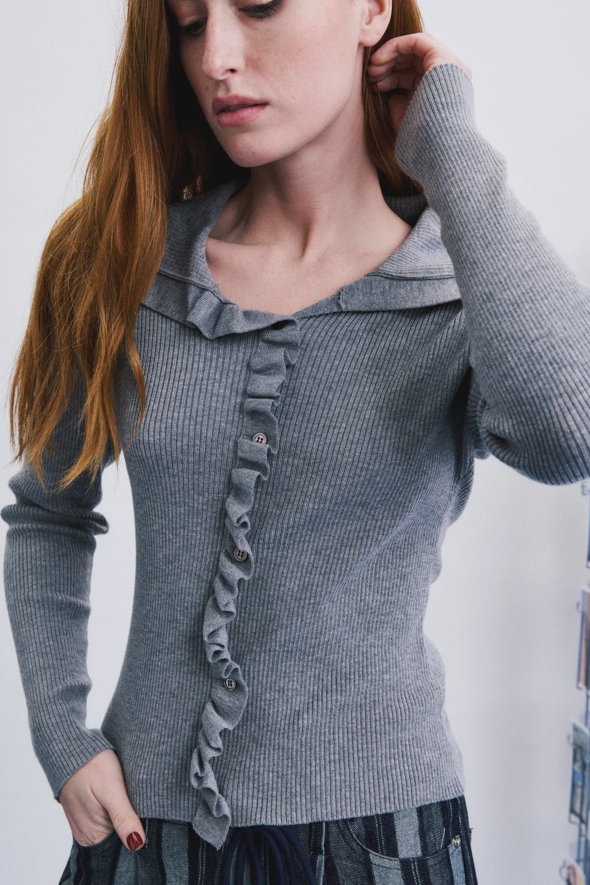 A woman wearing a gray ribbed knit top with a ruffled collar and ruffle trim down the front, paired with a dark patterned skirt. She is looking down and touching her hair against a plain light background.