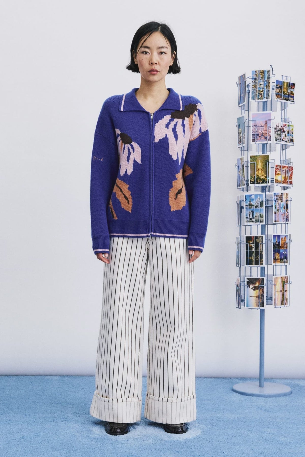 A model stands in a studio wearing a blue zip-up cardigan with abstract multicolor knit patterns and striped wide-leg pants. A clear acrylic photo display stand is visible to the right against a white wall and blue floor.