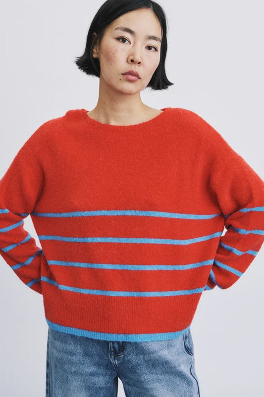 A model wearing a bright red knit sweater with horizontal light blue stripes across the chest and sleeves, paired with blue jeans, against a plain light gray background.