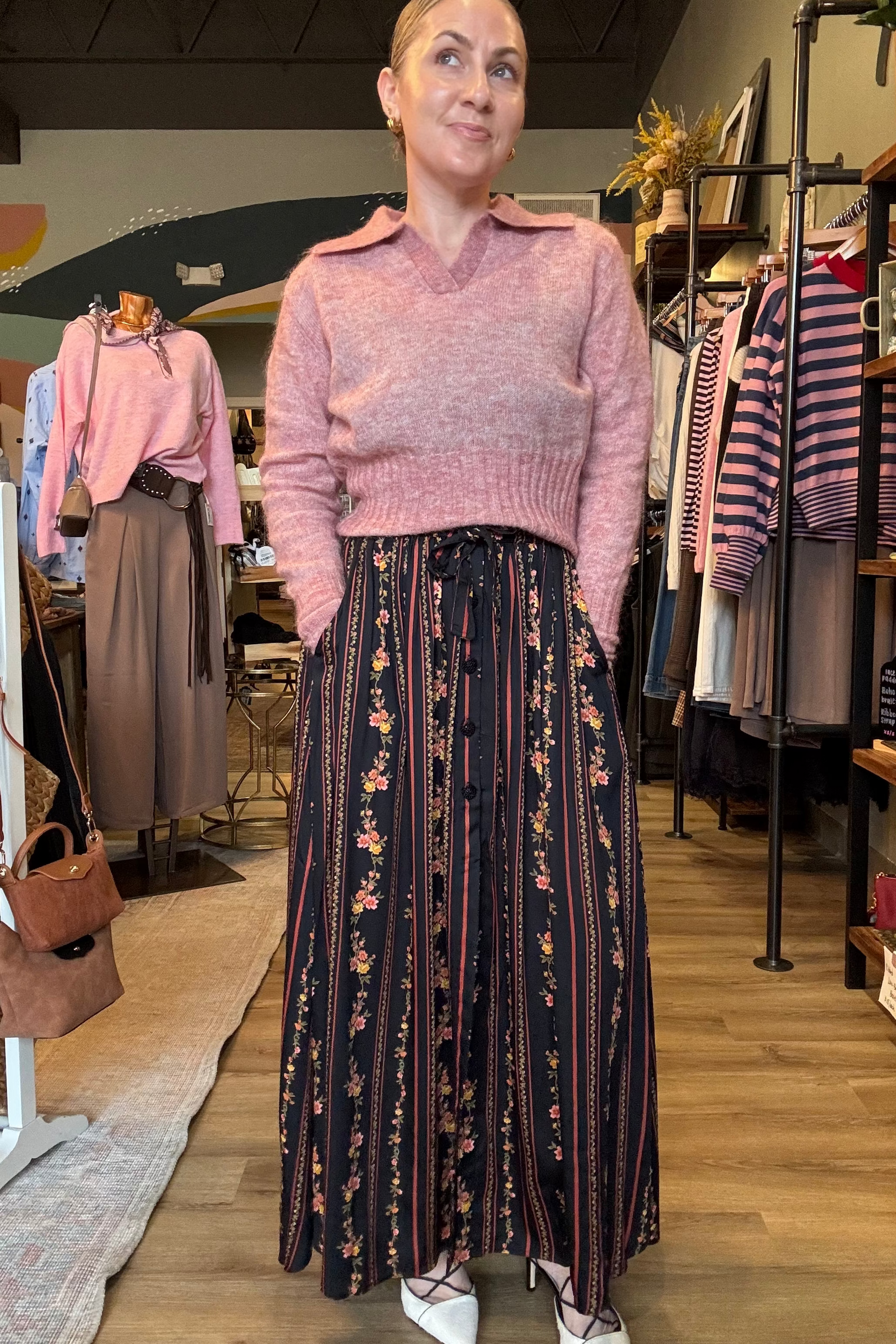 A woman stands inside a clothing boutique wearing a pink knit sweater with a collar and a long dark skirt with vertical stripes and a floral print. Clothing racks, shelves, and mannequins are visible in the background.