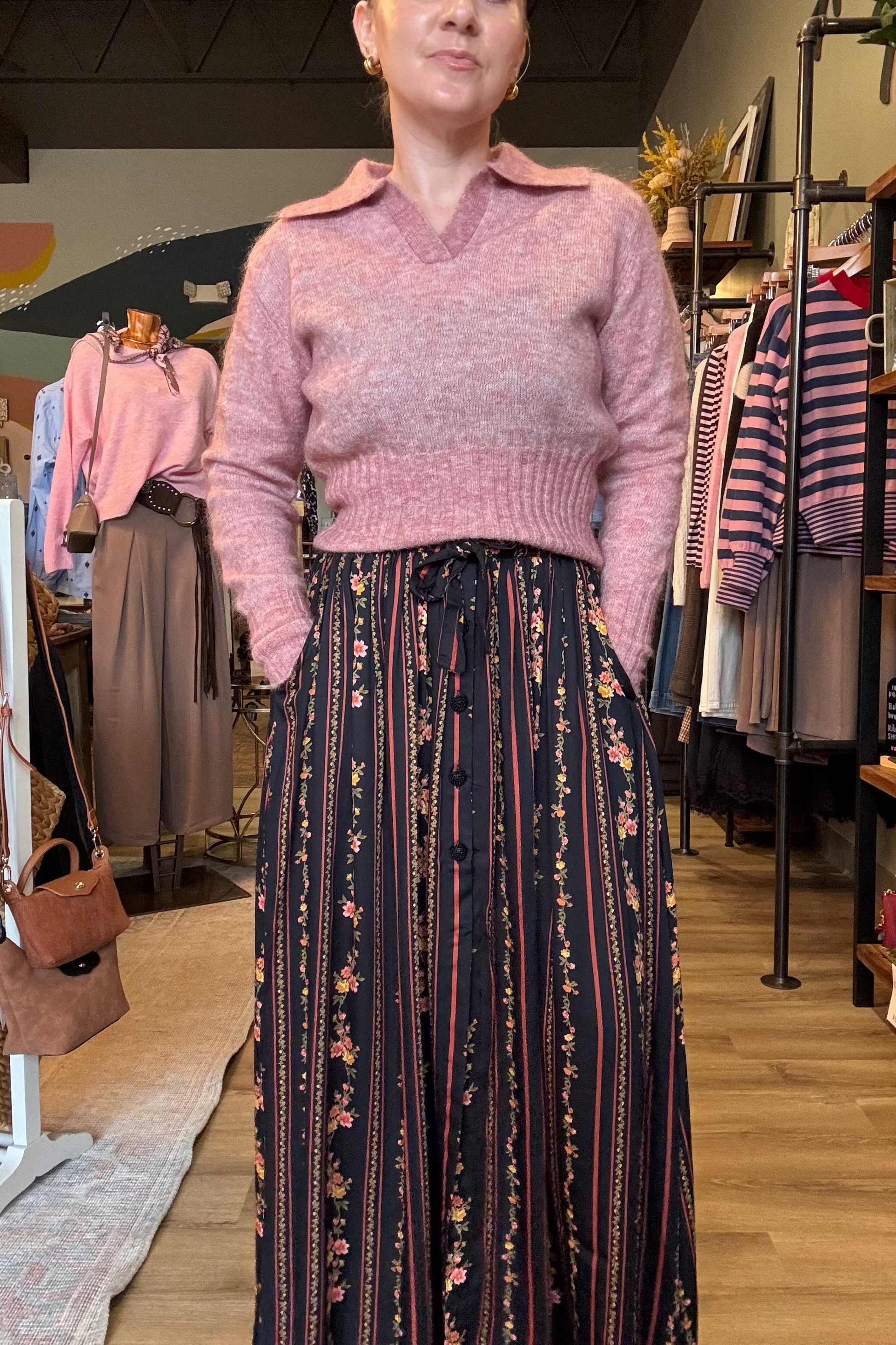 A woman modeling a dusty pink knit sweater vest with a collar over a long navy maxi skirt featuring vertical stripes and small floral motifs, standing inside a clothing boutique.