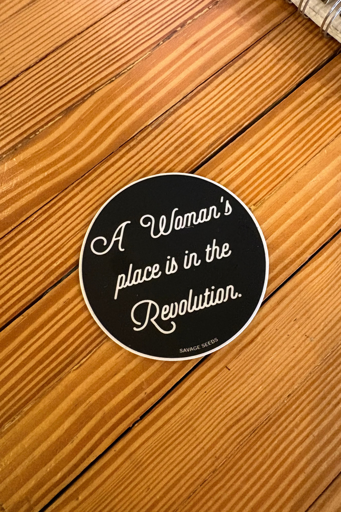 Round sticker with text 'A Woman's place is in the Revolution' on a wooden surface
