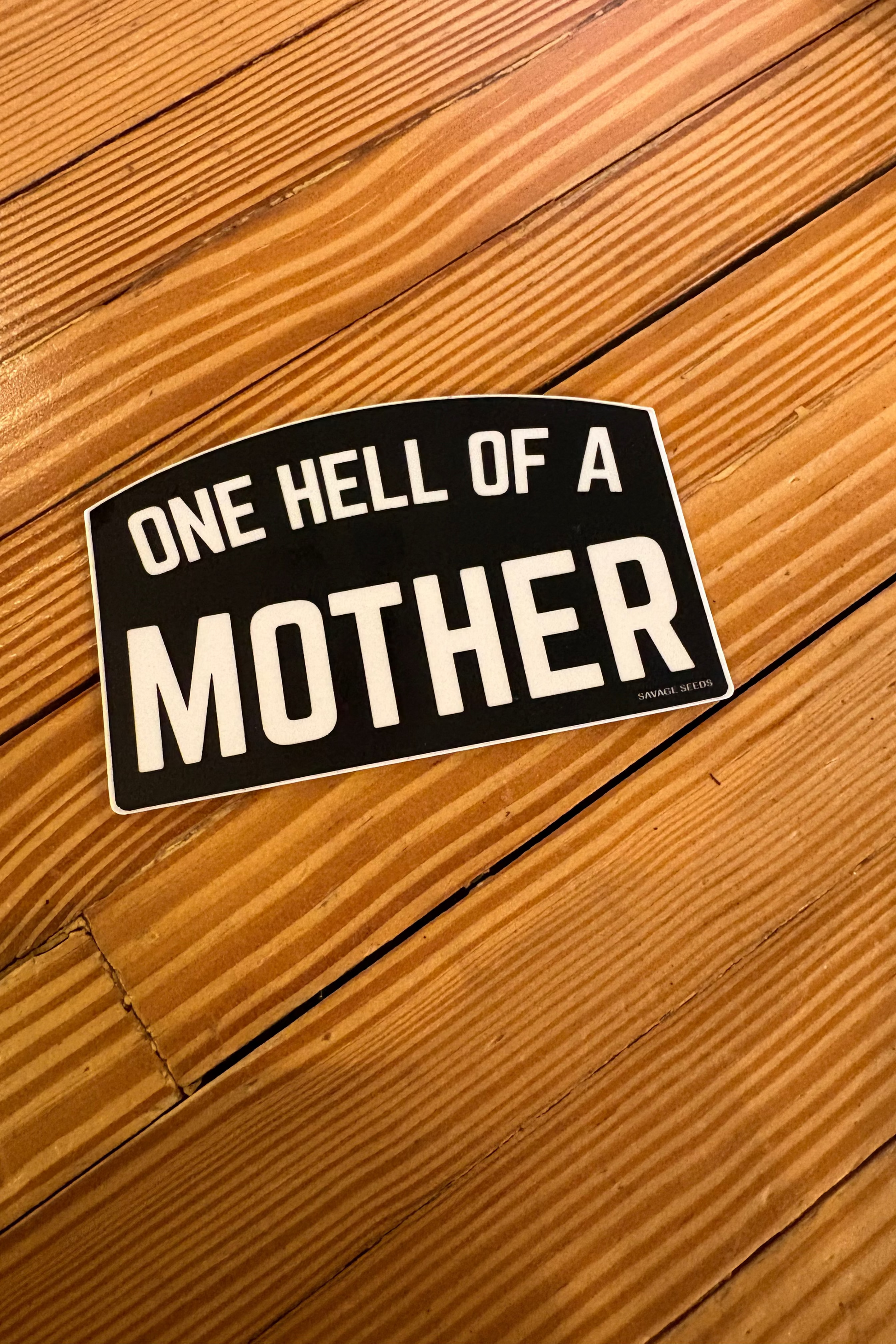 Sticker with 'One Hell of a Mother' text on a wooden surface